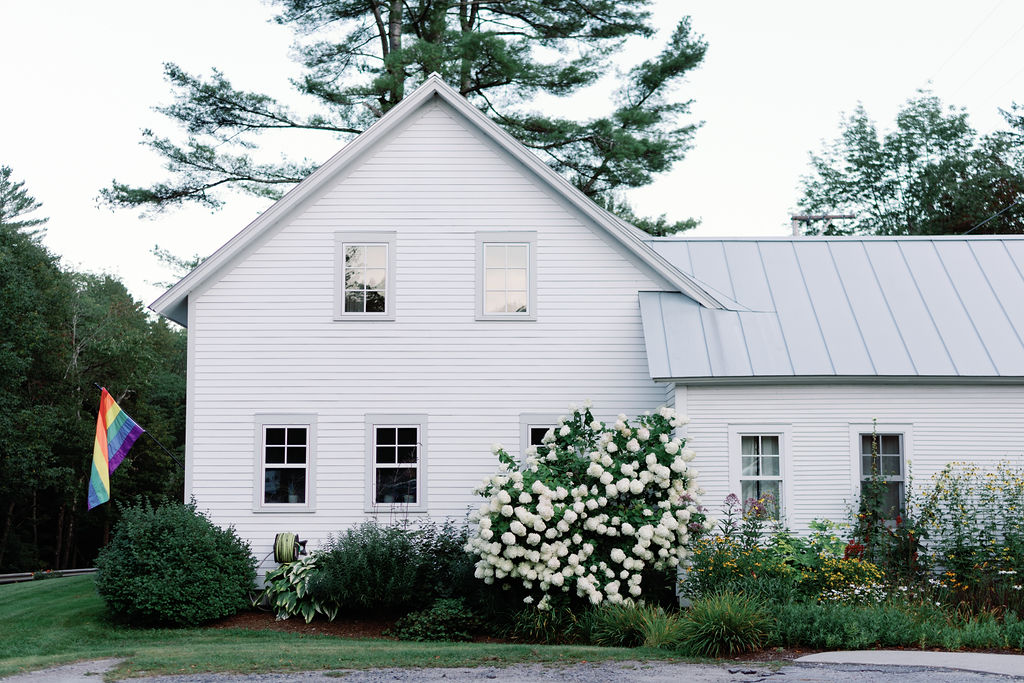 White farmhouse bearing a pride flag. Building is surrounded by a mature cottage garden bearing a large cloud white hydrangea and many other flower species.