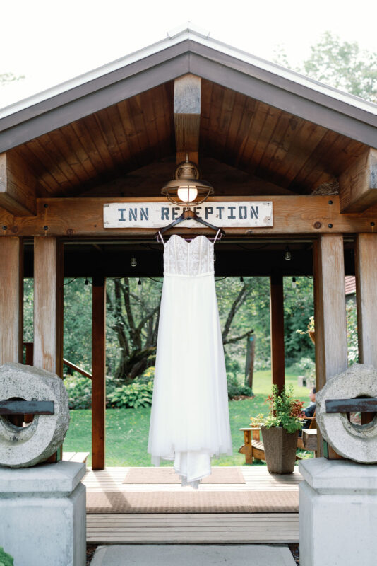 Rustic country breezeway with a a wedding dress hanging elegantly from its brass light fixture. There is a view of the lush gardens beyond.