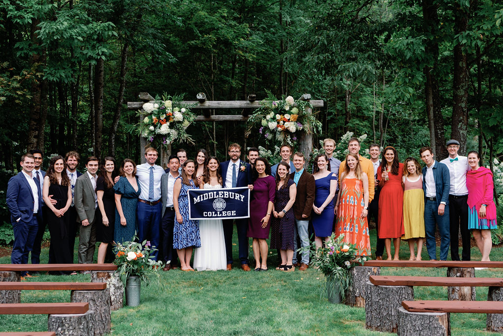 A group of people stand in front of a wooden archway decorated with flowers, while holding a banner that reads Middlebury College