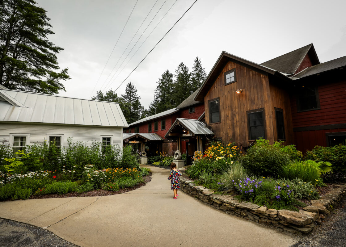 Entrance to the Mad River Barns restaurant/pub and reception area. Buildings are surrounded by verdant summer gardens. A young girl in a flowery sundress meanders down the main walkway to the buildings.