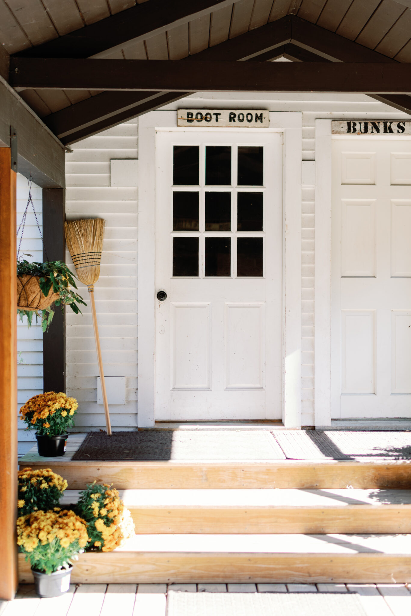 Side of our white farmhouse with two doors. The first has glass panes and a sign saying "boot room" above it, the second is all wood and bears a sign saying "bunks". The steps to the doors are lined with potted amber colored chrysanthemums.