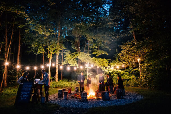 Night time around the fire pit. The area is surrounded by trees strung together with lights. Guests are enjoying the smores bar.