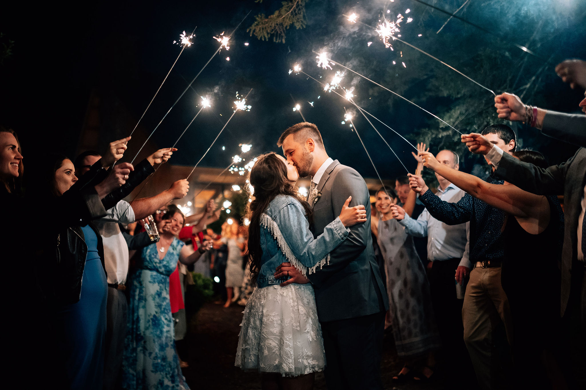 Bride and groom kiss underneath a tunnel of sparklers created by their guests before ending the night. Bride is wearing short white lace dress and denim jacket with beaded fringe. Groom is in grey suit.