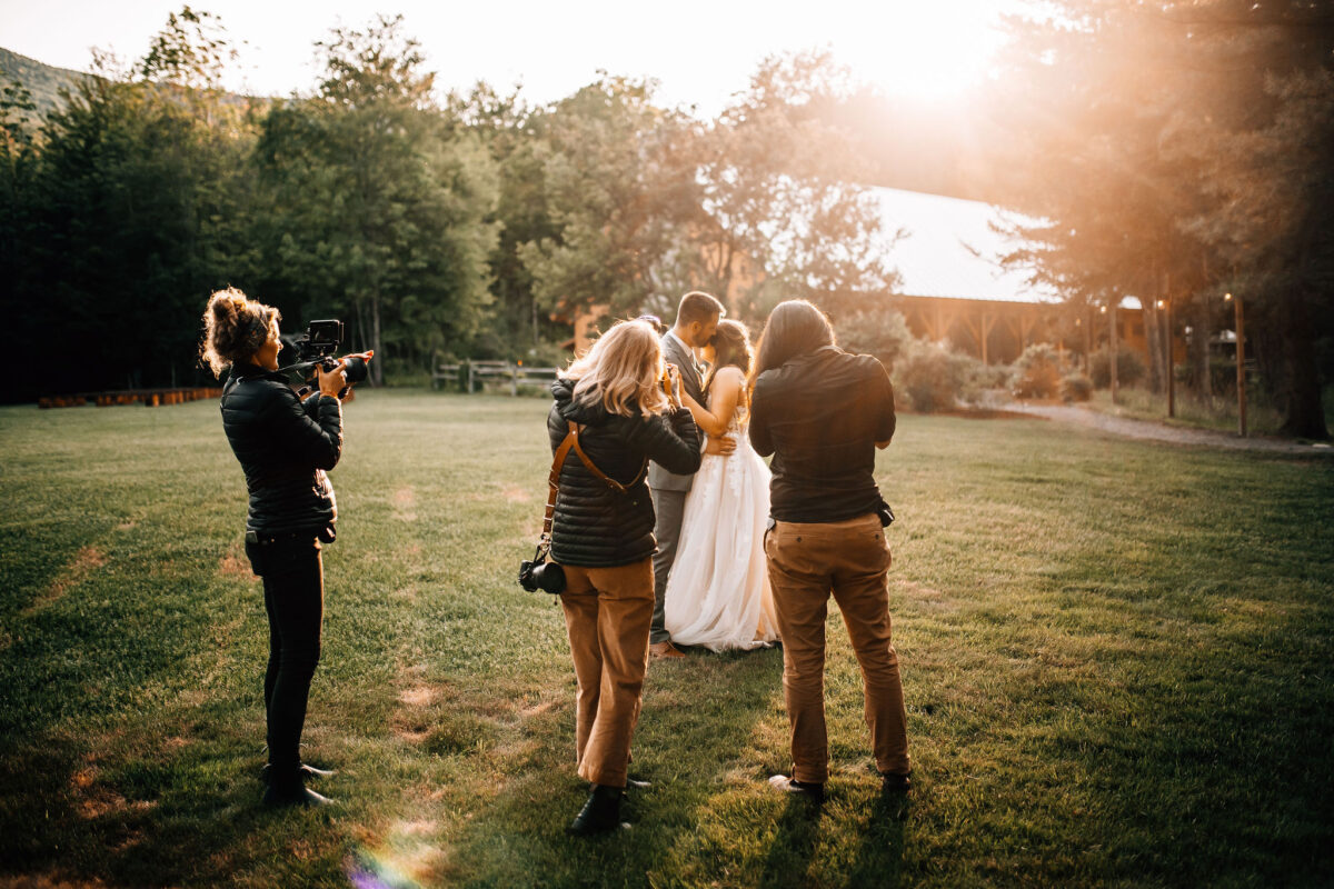 Bride and Groom embracing in a sunny field as photographers and videographers capture the moment.